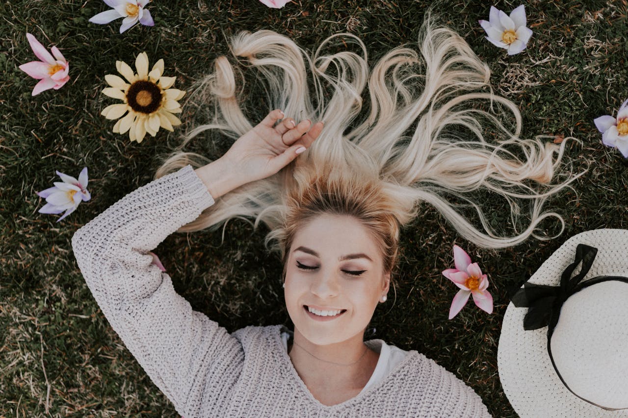 about-img A smiling woman with closed eyes lies on grass, surrounded by flowers, capturing a moment of pure relaxation.