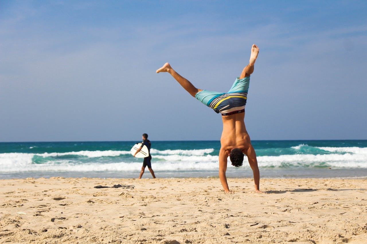 our-experience Man doing a handstand on a sunny beach with surfer in background, evoking a sense of freedom and joy.