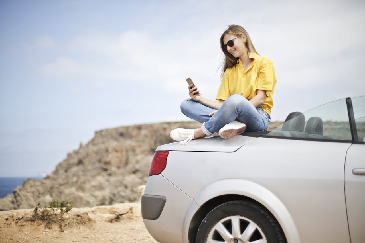 ours-journey Smiling young woman sitting on a car at the beach in Mgarr, Malta using a smartphone.